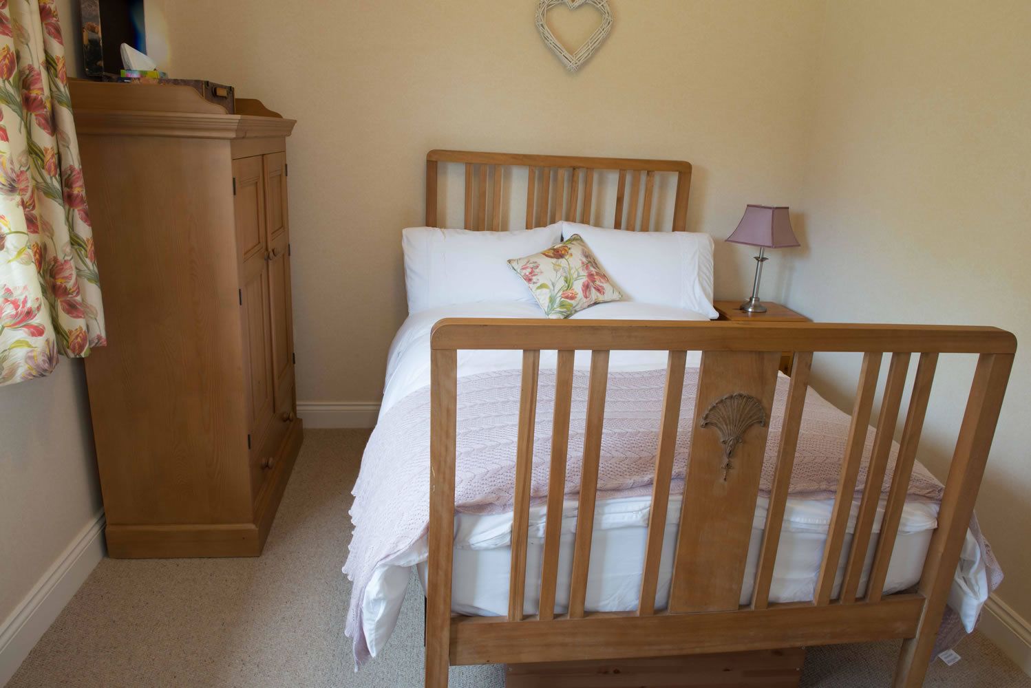 A bedroom with a wooden bed, a nightstand, a lamp, a wardrobe, and a heart-shaped decoration on the wall.