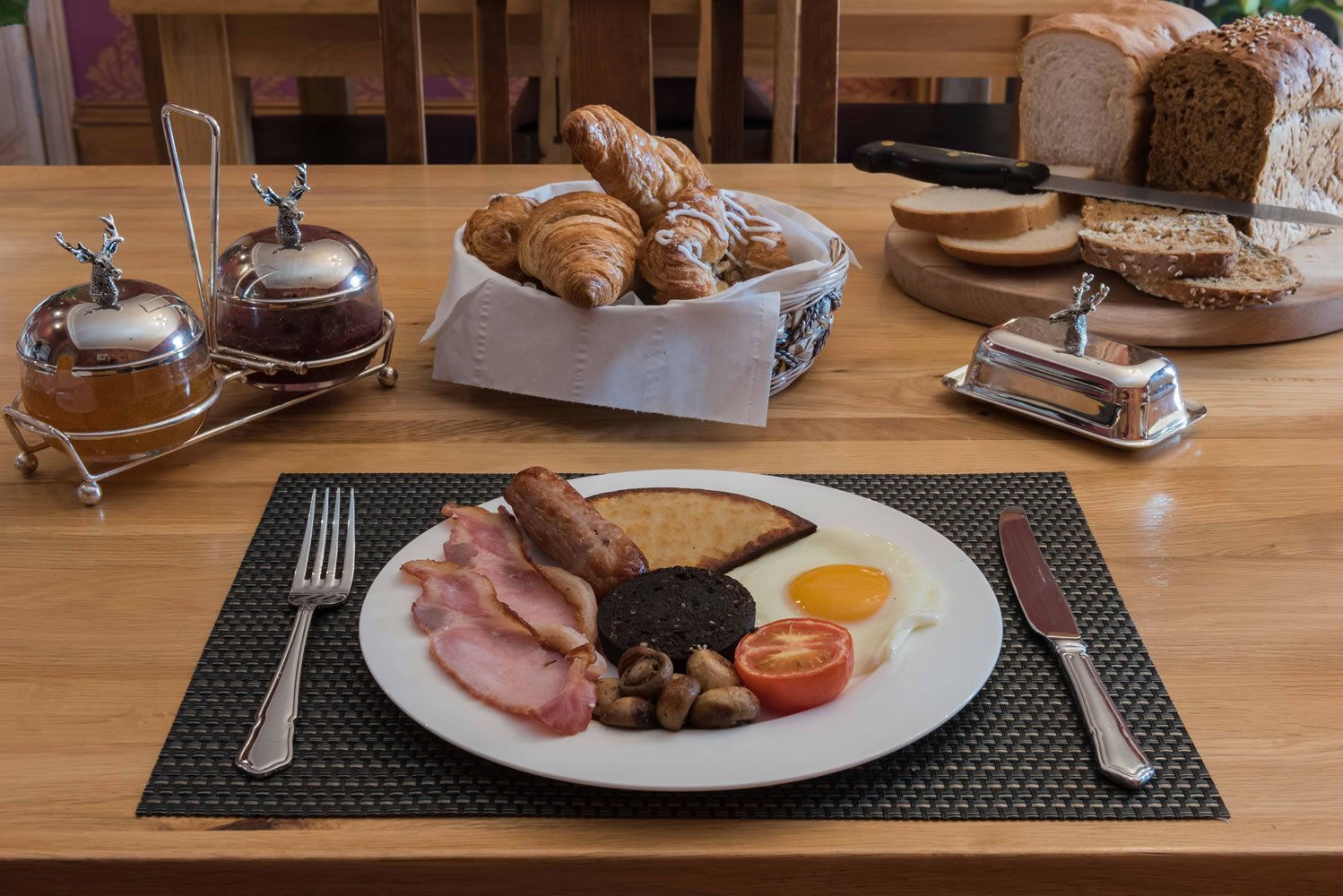 Une table de petit-déjeuner avec des croissants, du pain, de la confiture et une assiette contenant du bacon, des œufs, des champignons et une tomate.