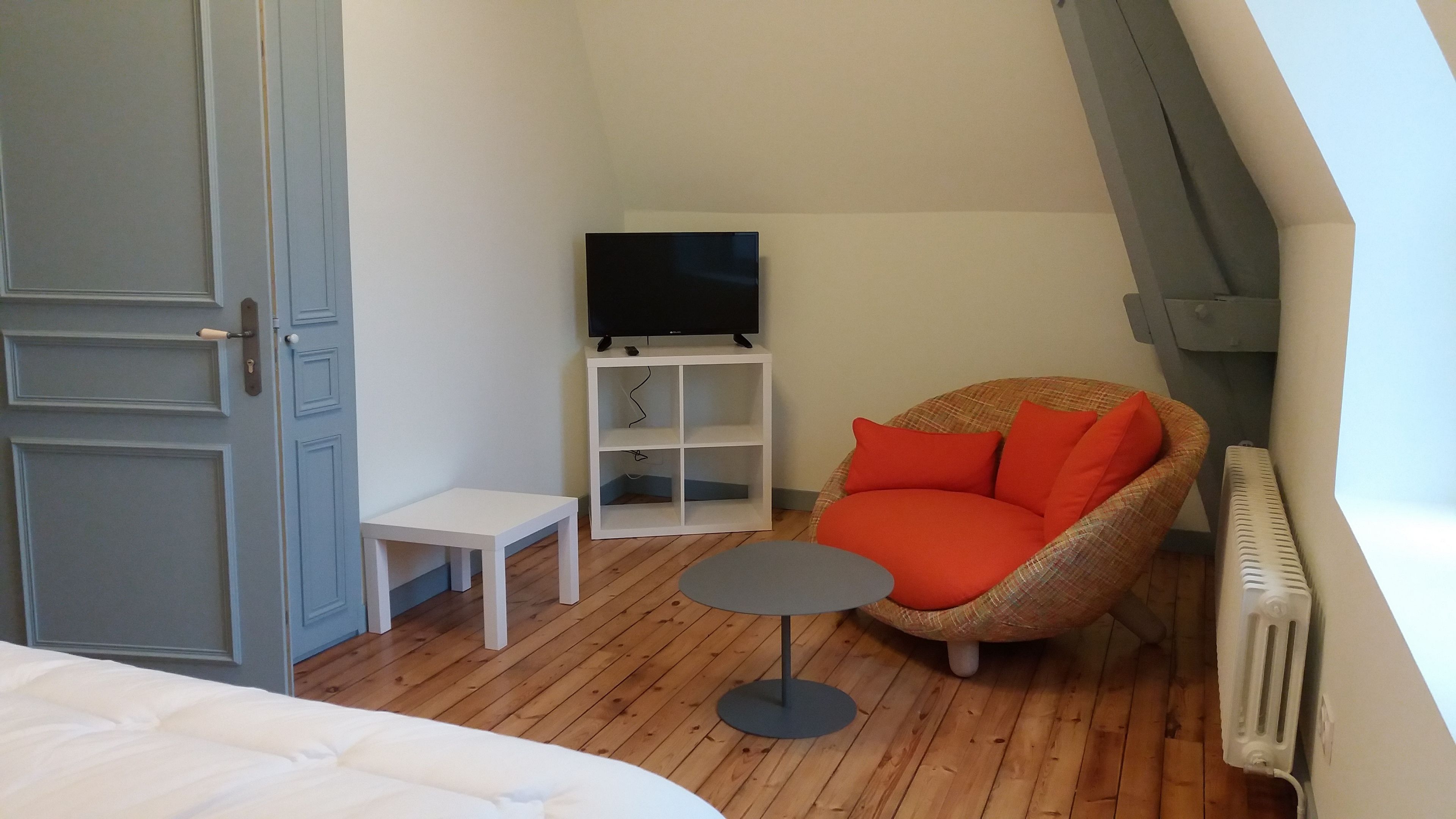 A small, cozy room with a bed, a chair, a small table, a TV on a white stand, and a radiator.