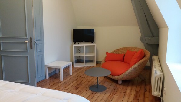 Una habitación pequeña y acogedora con cama, una silla, una mesita, televisor sobre un soporte blanco y un radiador.