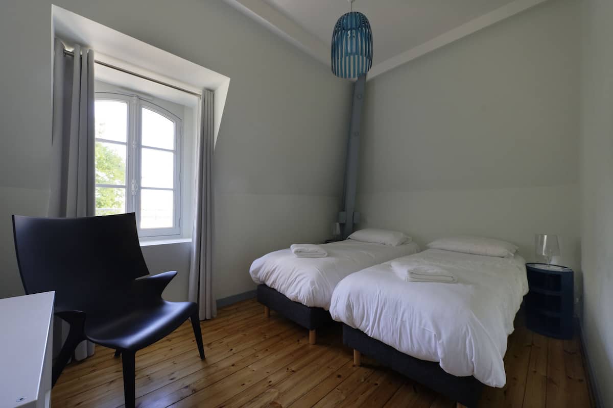 A bedroom with two beds, a chair, a desk, and a window with curtains.