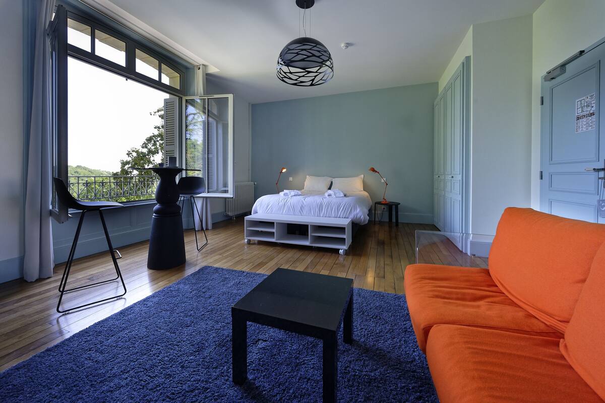 A bedroom with a bed, a black table, an orange couch, and a large window.