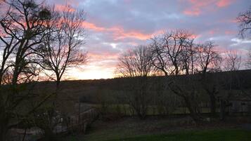 Un coucher de soleil sur un paysage rural, avec des arbres sans feuilles et un bâtiment situé au loin.