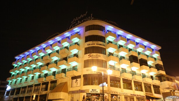 Un hôtel à plusieurs étages, doté de balcons illuminés et d’enseignes indiquant « Hôtel Grand Gabriel » et « Salon de coiffure ».