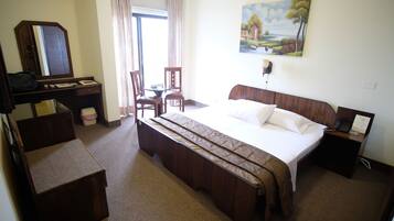 A hotel room with a bed, a desk with a chair, a wardrobe, and a painting on the wall.