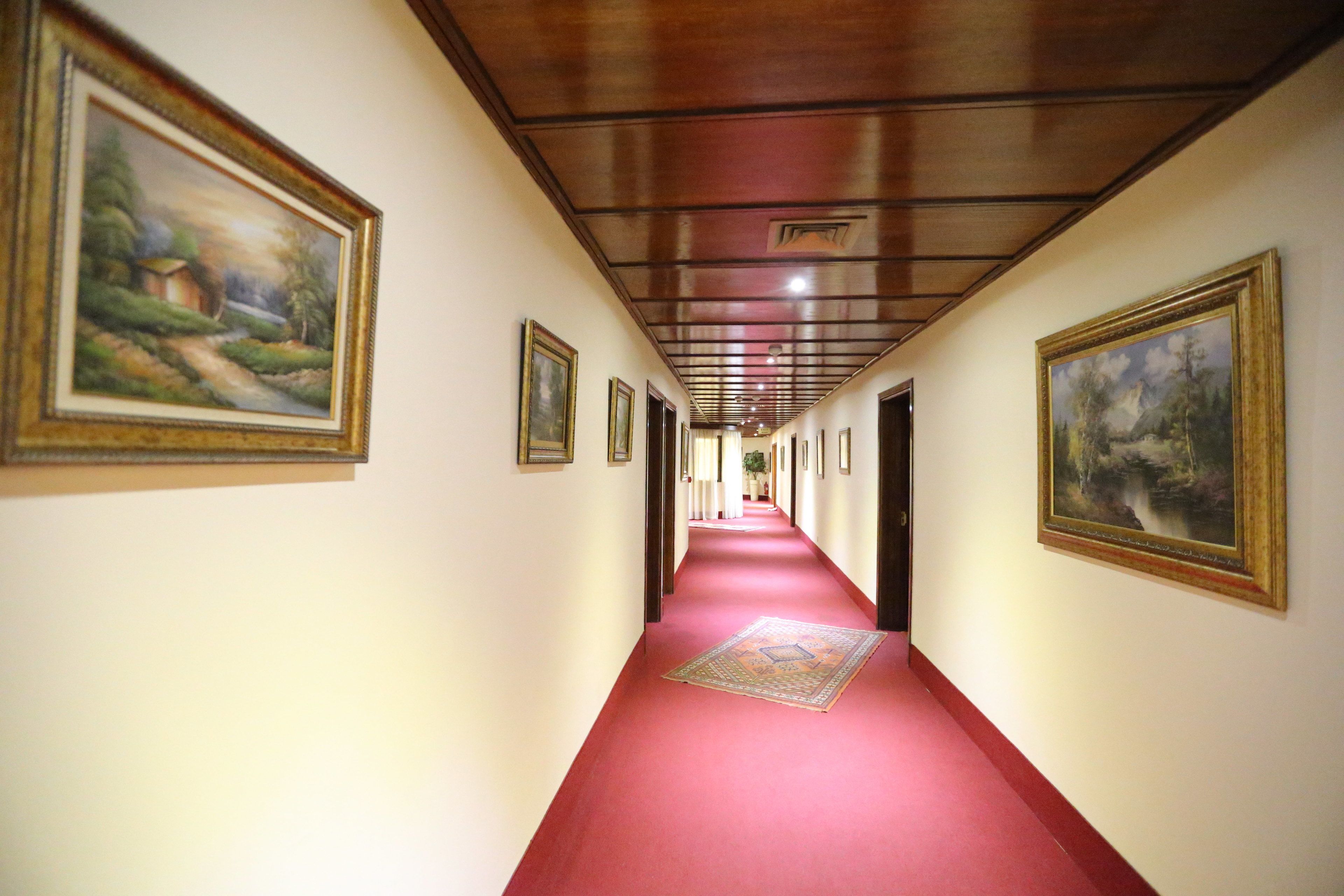 A hallway with a red carpet, wooden ceiling, and framed paintings on the walls.