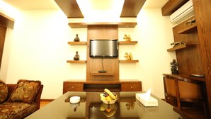 Un salon moderne avec un téléviseur à écran plat fixé à un mur en bois, une table basse en verre sur laquelle repose un panier de fruits, et un canapé agrémenté de coussins à motifs.
