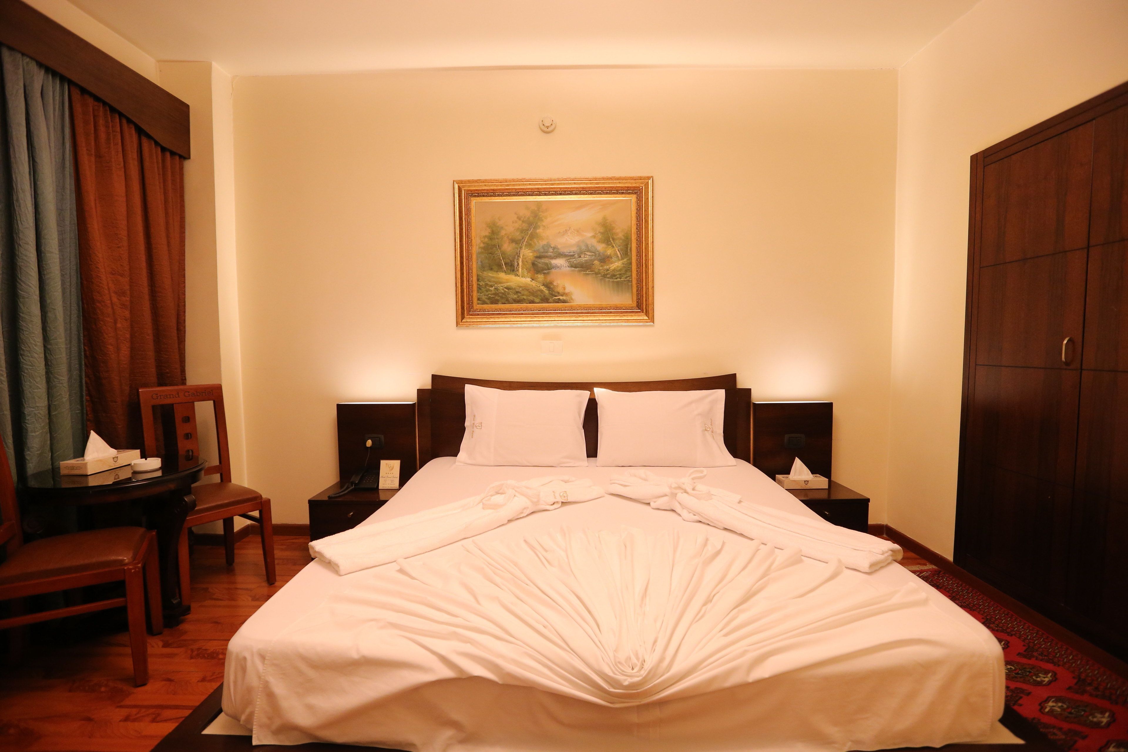 A neatly made bed with white linens, a wooden headboard, and two bedside tables with lamps. A painting of a landscape hangs above the bed.