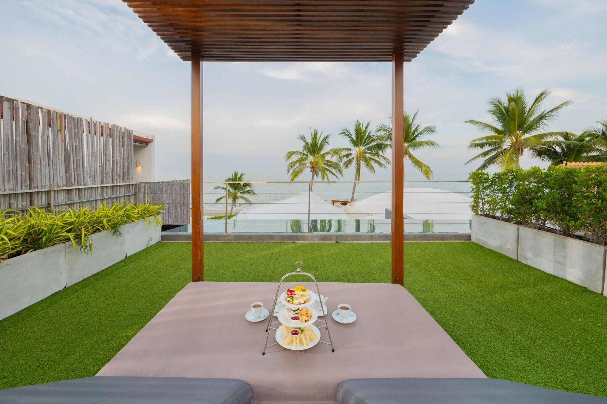 A terrace with a three-tiered outdoor dining set, a wooden pergola, and a view of the sea and palm trees.