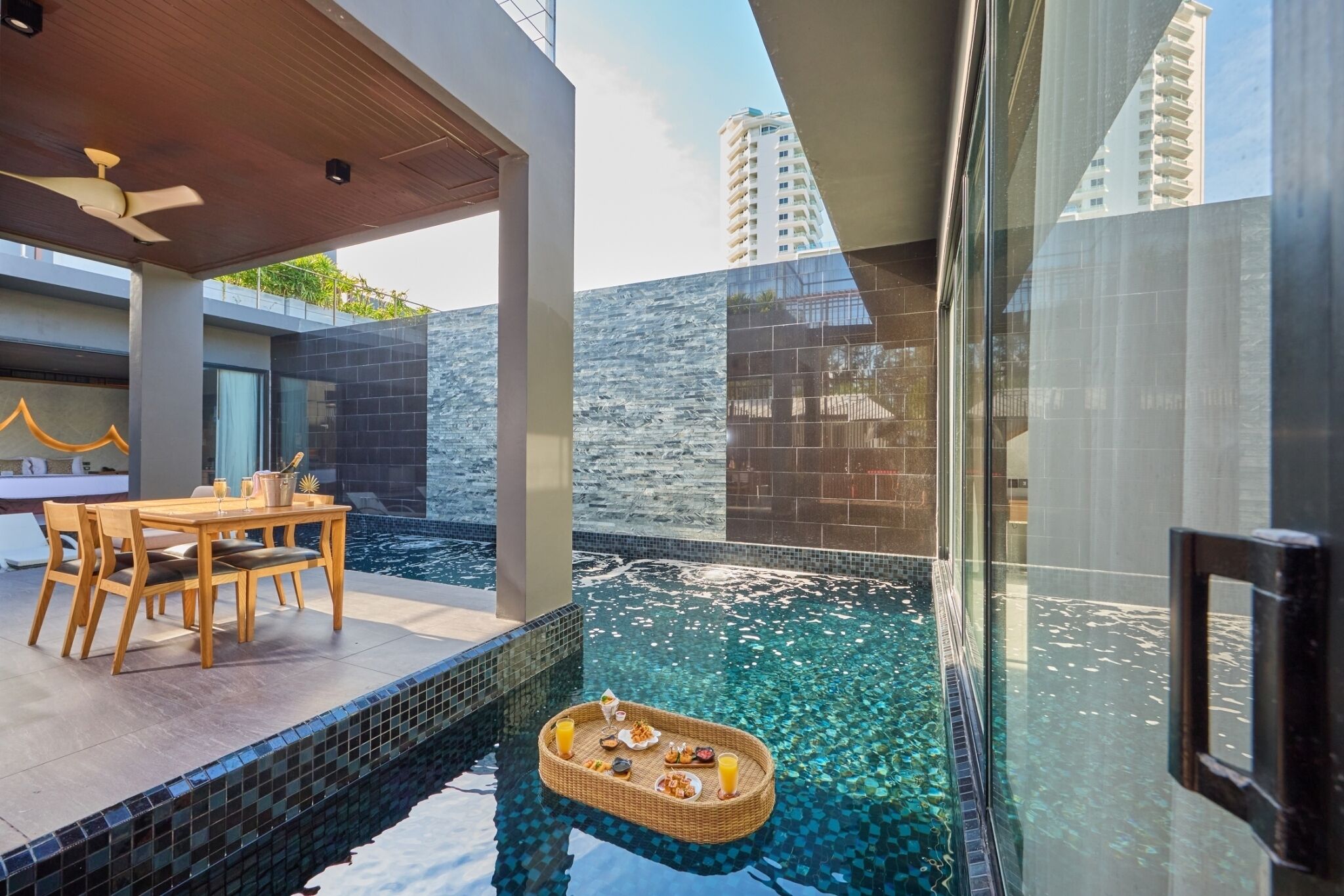 Un espace piscine extérieur moderne, doté d’une table et de chaises, d’un plateau flottant avec de la nourriture et des boissons, et offrant une vue sur le paysage urbain.