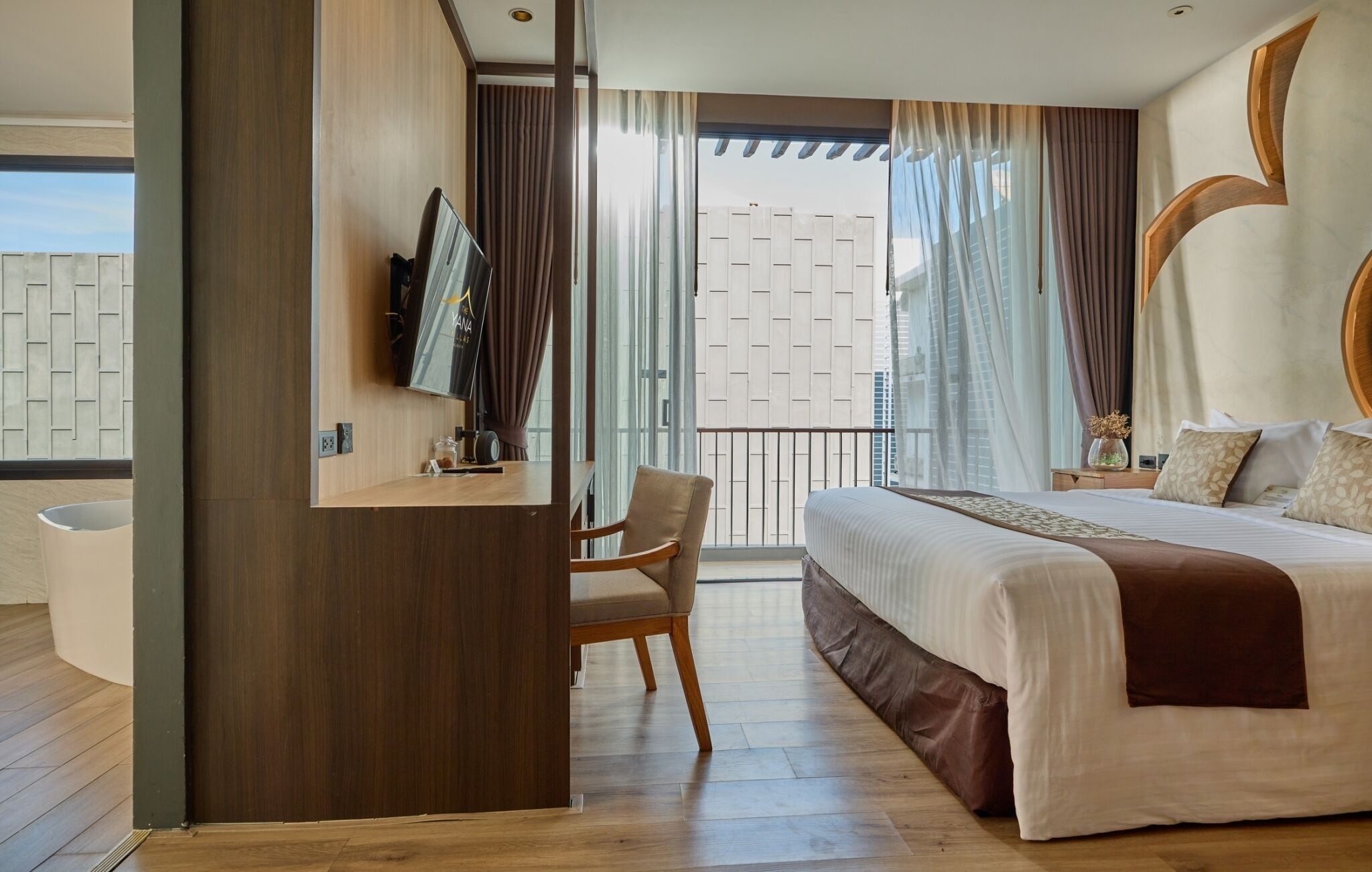 Ett modernt hotellrum med en stor säng, ett skrivbord med en platt-TV, en stol och en balkong med gardiner.