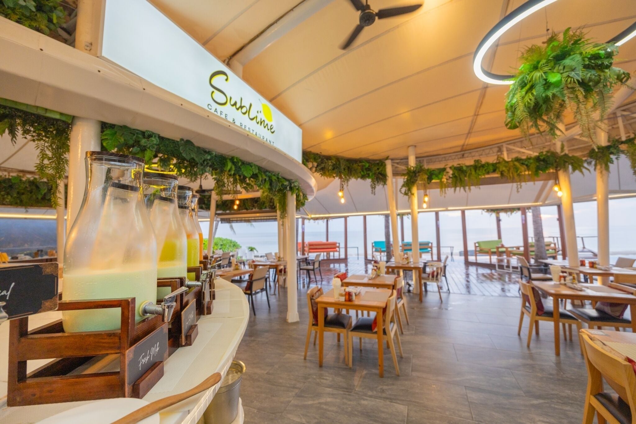 A restaurant with a high ceiling, hanging plants, and a view of the sea.