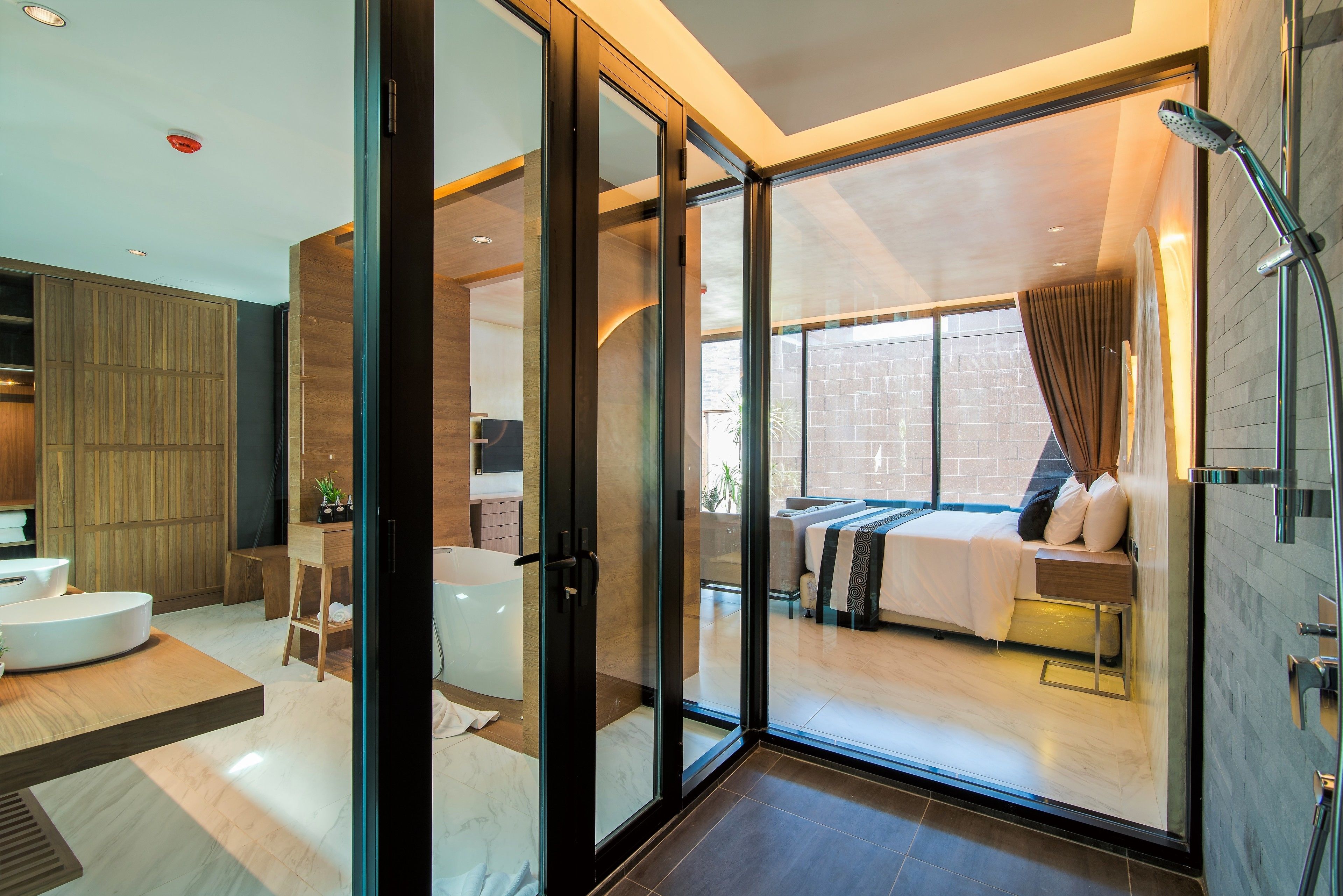 A modern hotel room with a glass door leading to a balcony, a large bed, a separate bathroom, and a round white bathtub.