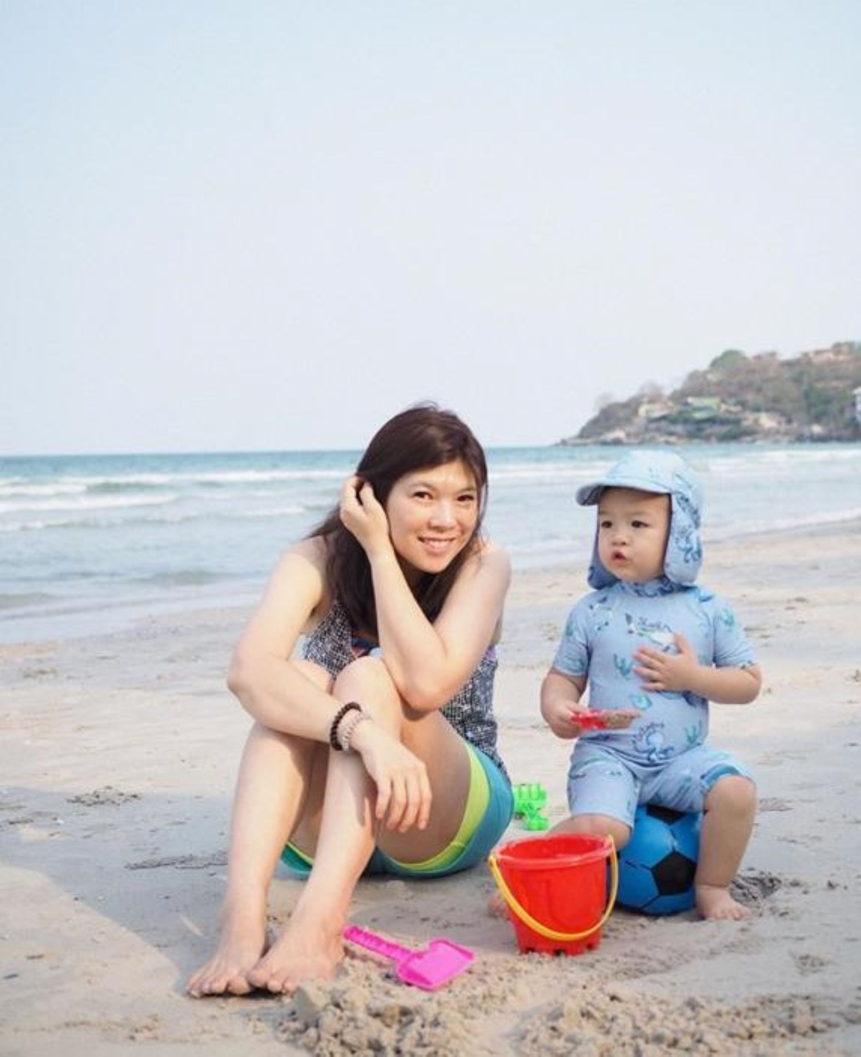 A beach scene with a person sitting and a child playing with a ball.