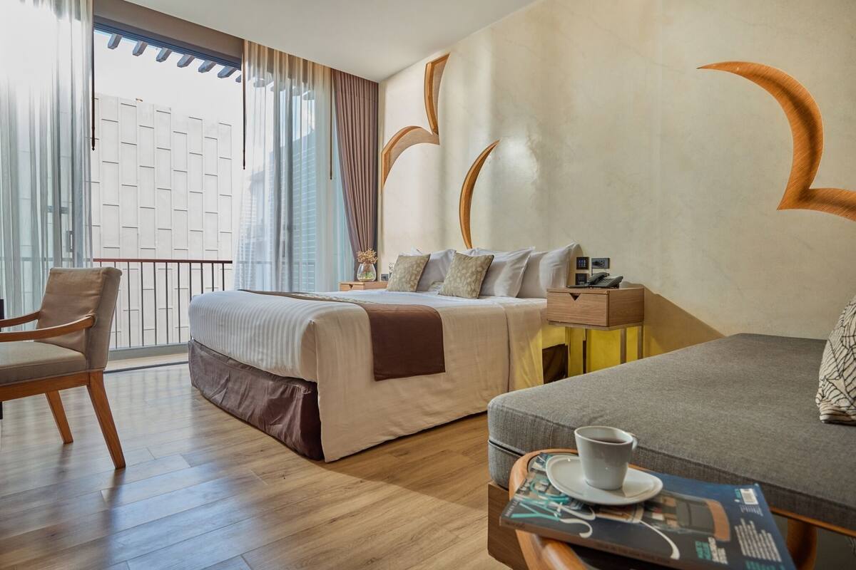Ein Hotelzimmer mit einem Bett, einem Sofa, einem Sessel, einem kleinen Tisch mit Tasse und Zeitschrift sowie einem Fenster mit Vorhängen.