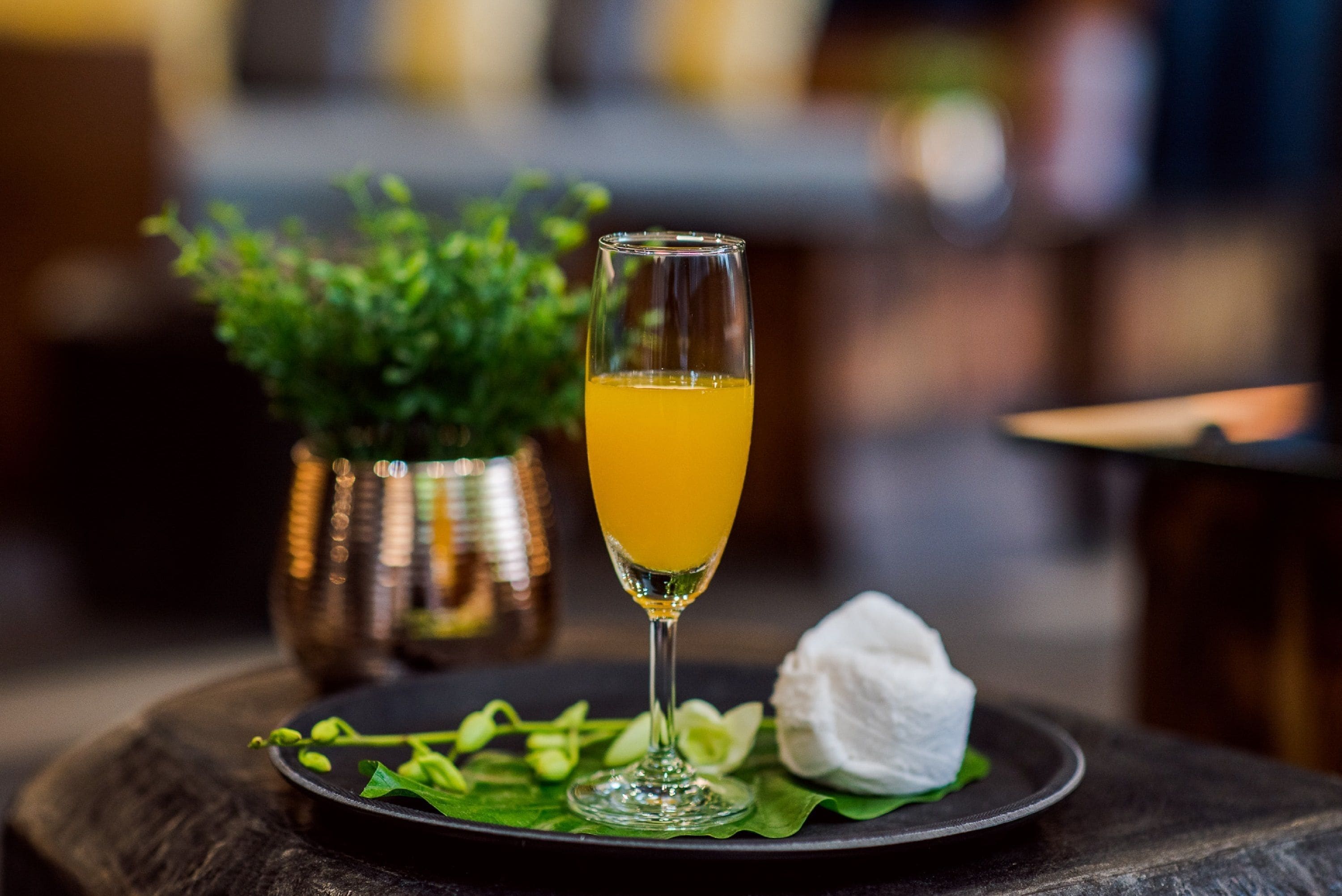 Een glas sinaasappelsap op een bord met groene bladeren en een wit voorwerp, mogelijk een bloem, op een houten tafel.