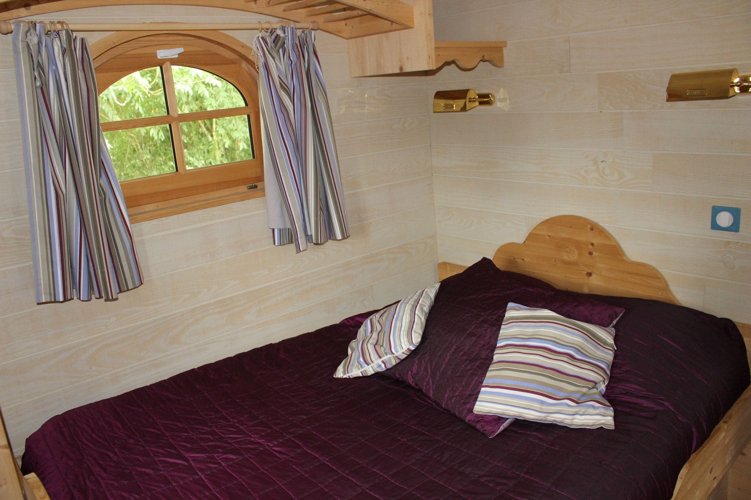 A wooden bed with a purple quilt and striped pillows, a window with curtains, and a wooden headboard.