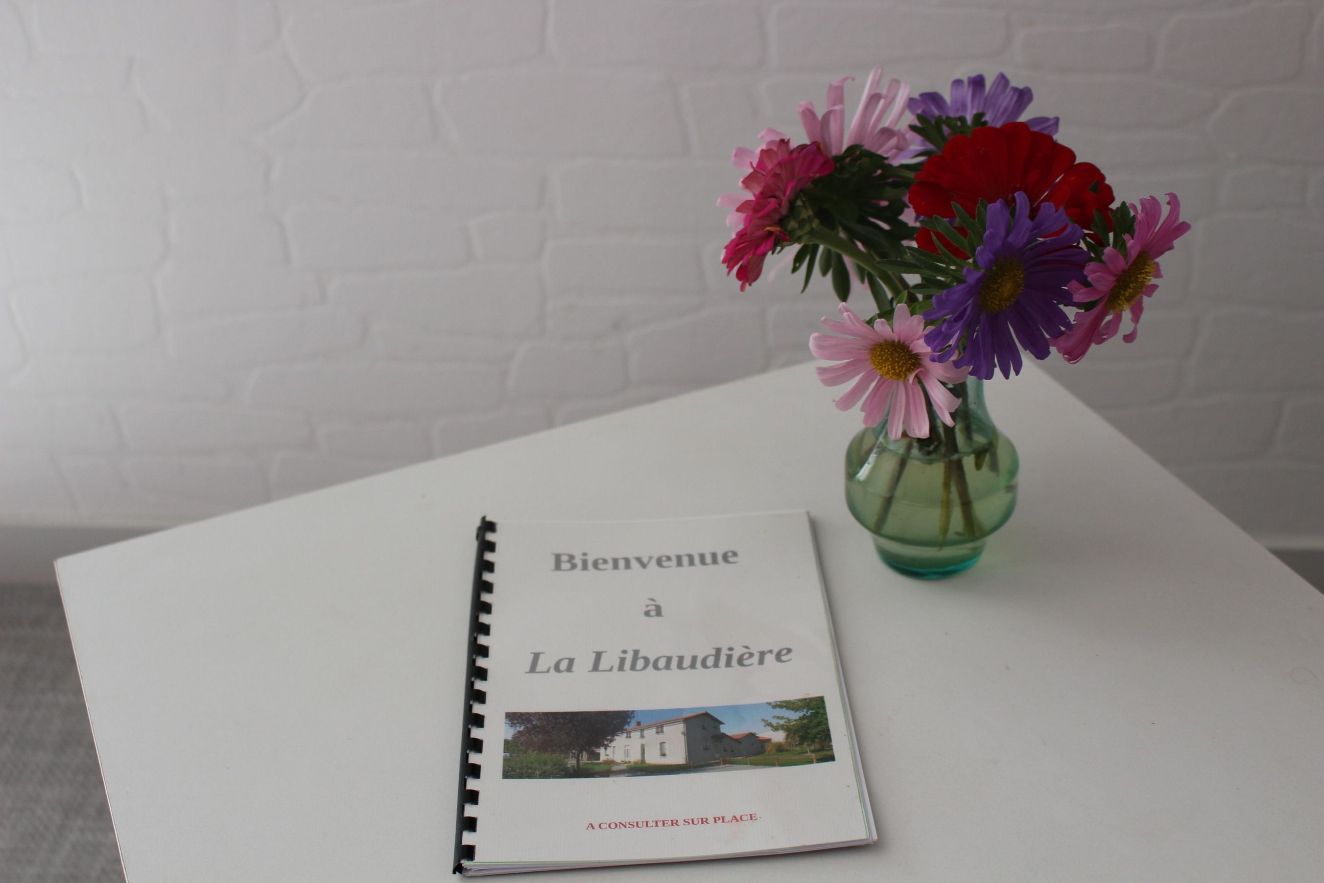 Un libro titulado “Bienvenue a La Libaudière” junto a un jarrón con flores sobre una mesa blanca frente a una pared de ladrillo blanco.
