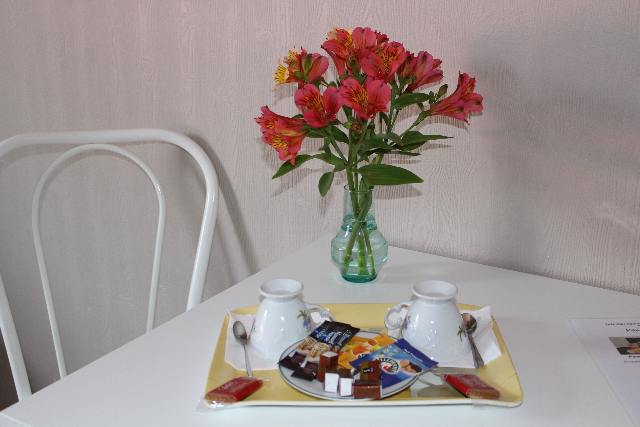 Una mesa con una bandeja de comida, dos tazas blancas, un jarrón verde con flores y una silla blanca.