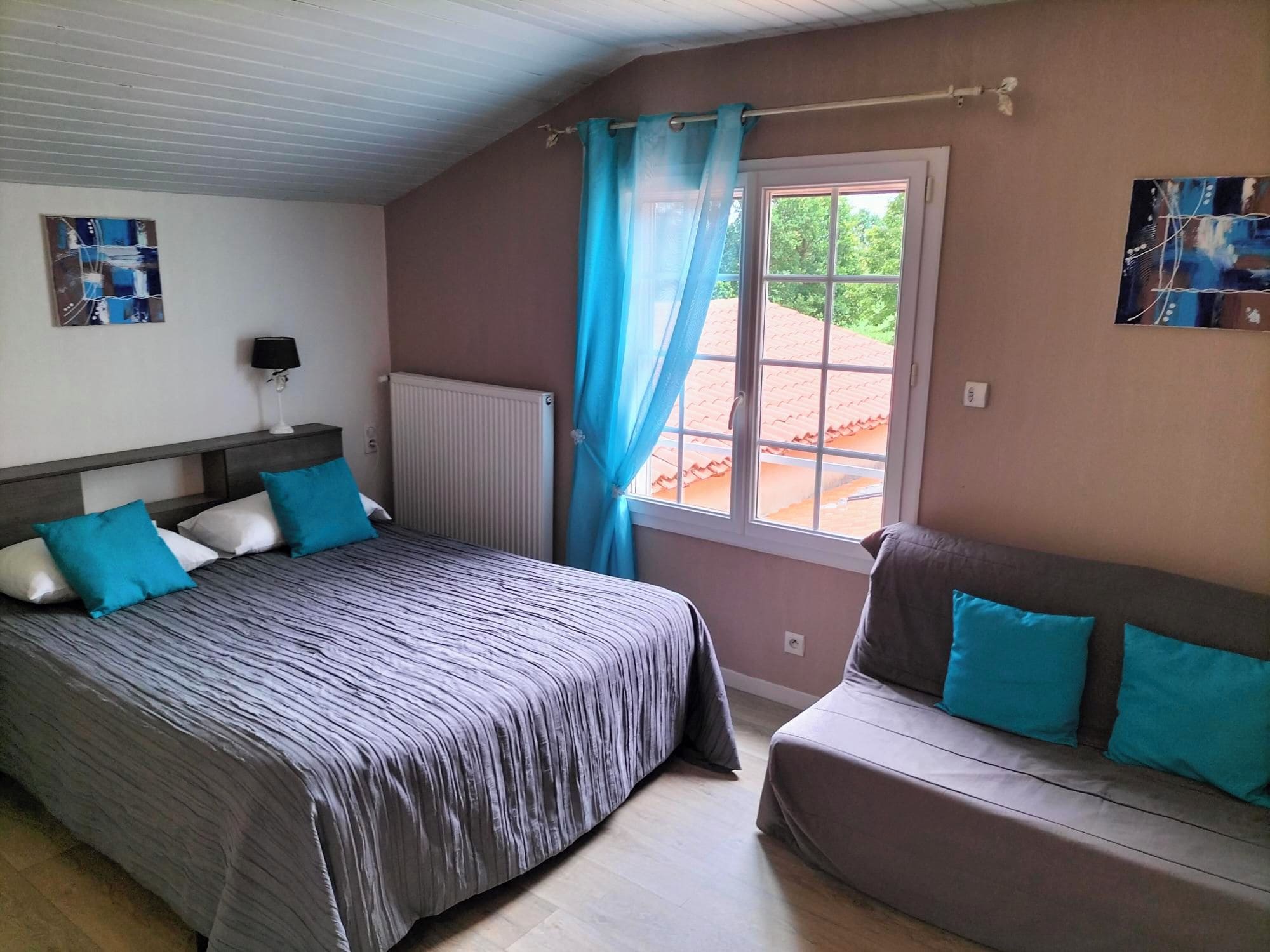 A bedroom with a bed, a sofa, a window with curtains, a radiator, and artwork on the walls.