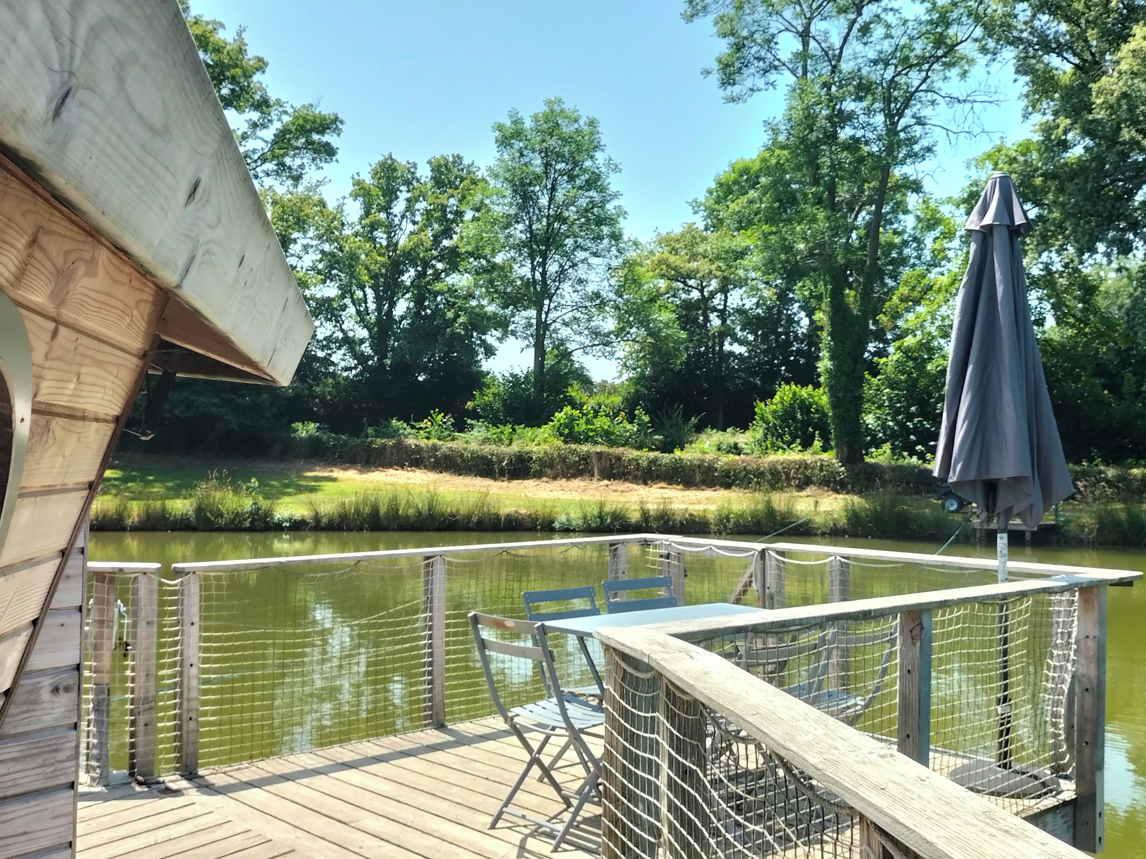 A wooden deck with a table and chairs, overlooking a calm body of water and surrounded by greenery.
