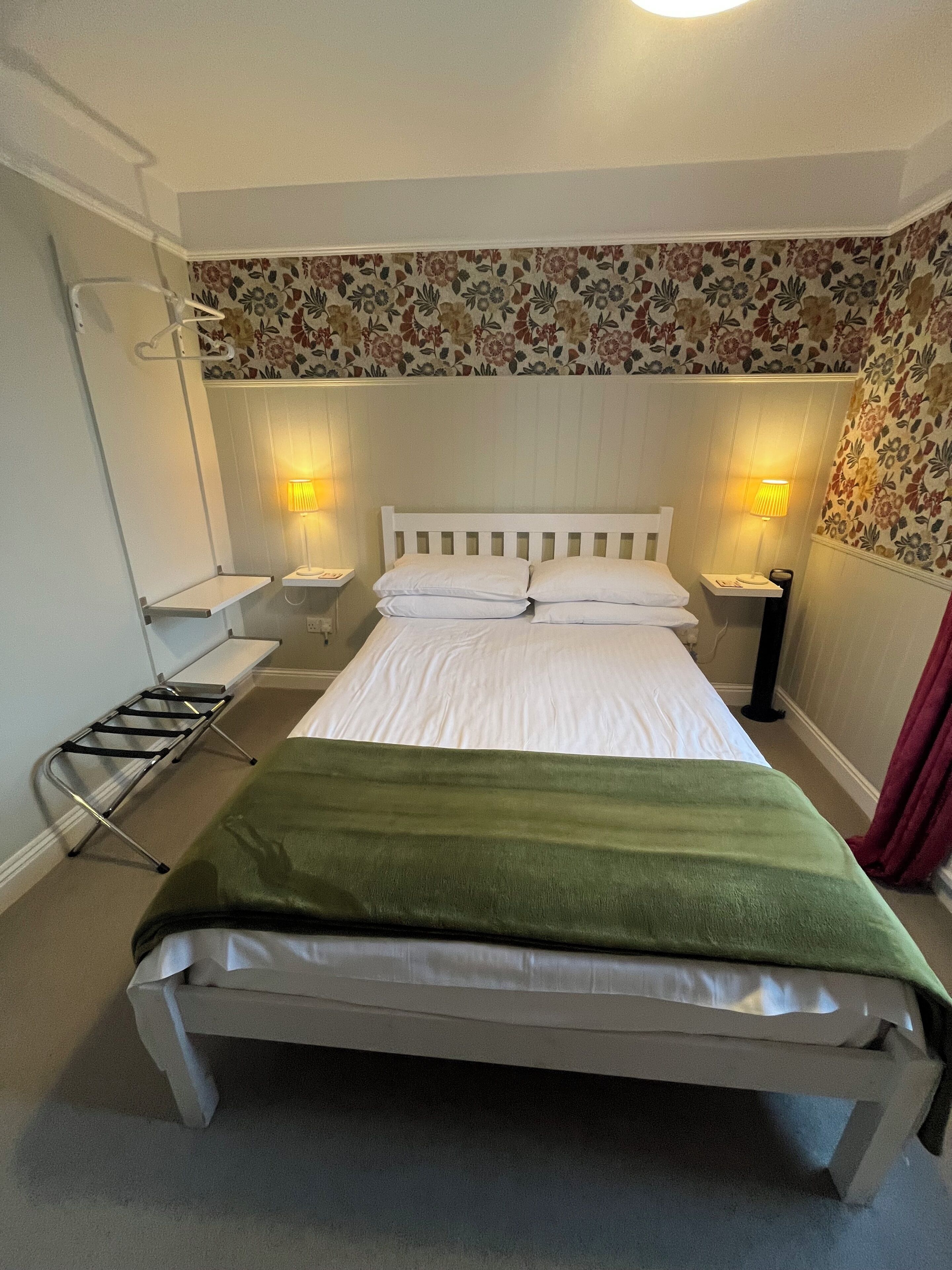 A neatly made bed with a green blanket, two bedside tables with lamps, and floral wallpaper.