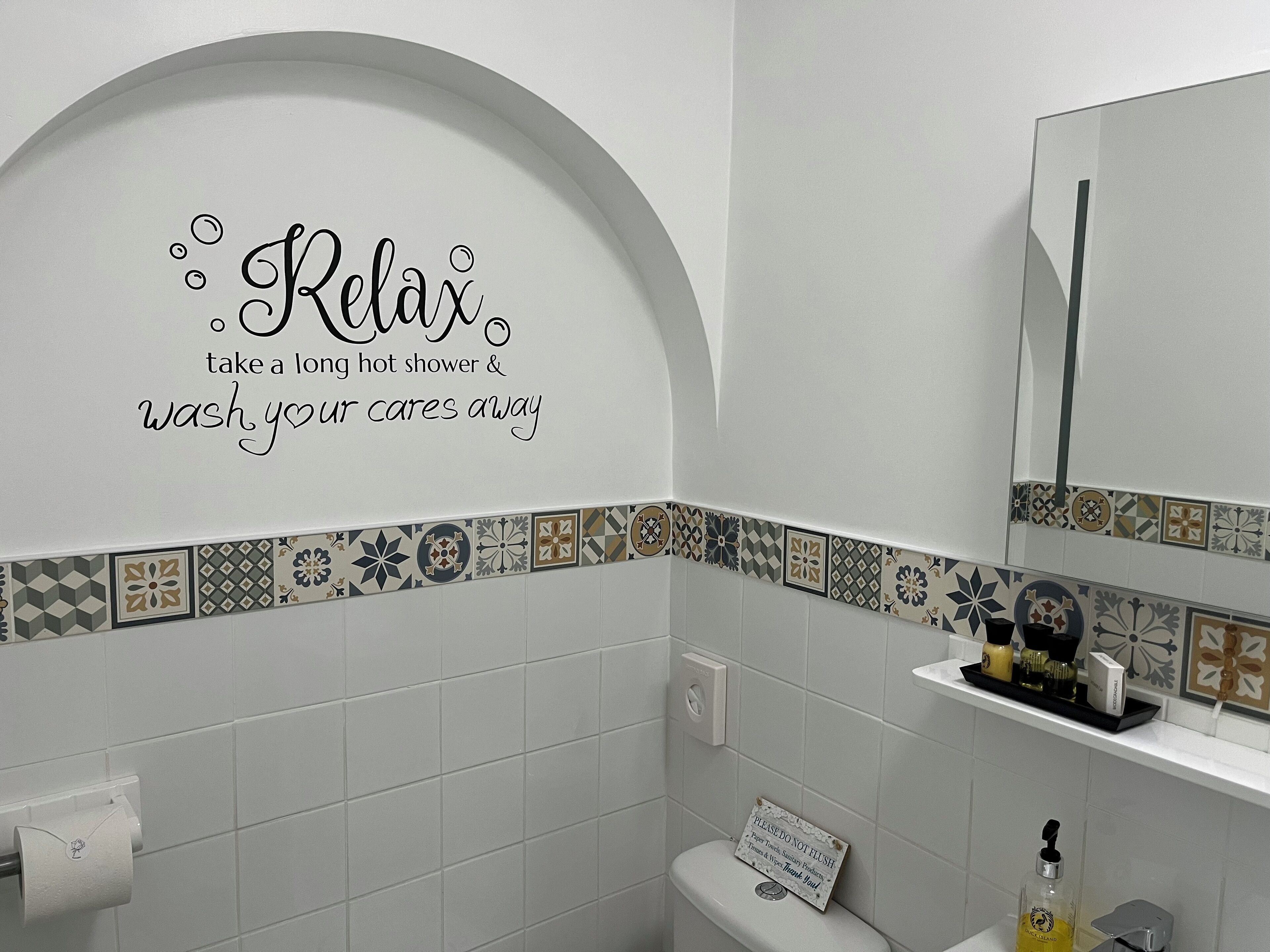 Une salle de bain avec un décor de bordure en carreaux, des toilettes, un lavabo avec des articles de toilette et un miroir.