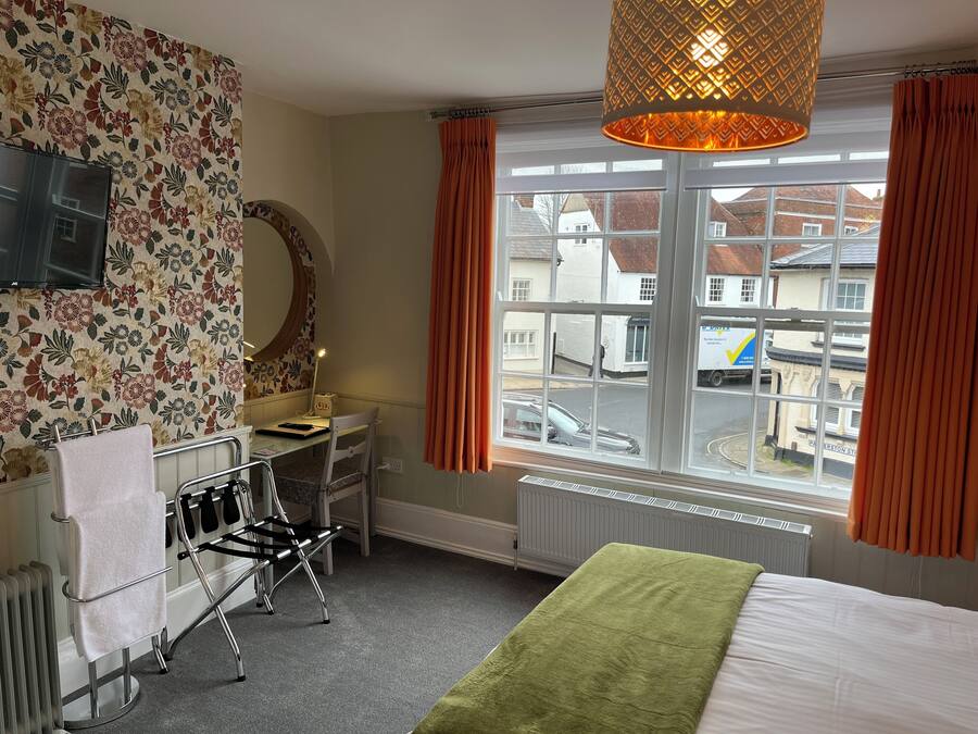 A hotel room with a bed, a desk with a chair, a large mirror, a radiator, and a window with curtains.