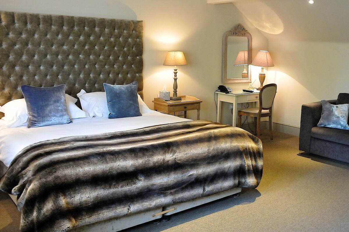 A hotel room with a tufted headboard, a bed with a dark patterned bedspread, a desk with a lamp, a chair, and a couch.