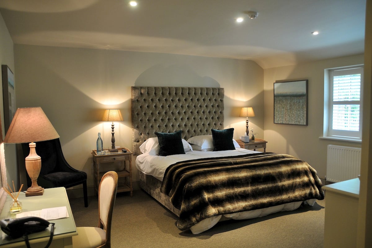 A hotel room with a large bed, two bedside tables with lamps, a chair, a desk, and a window with blinds.