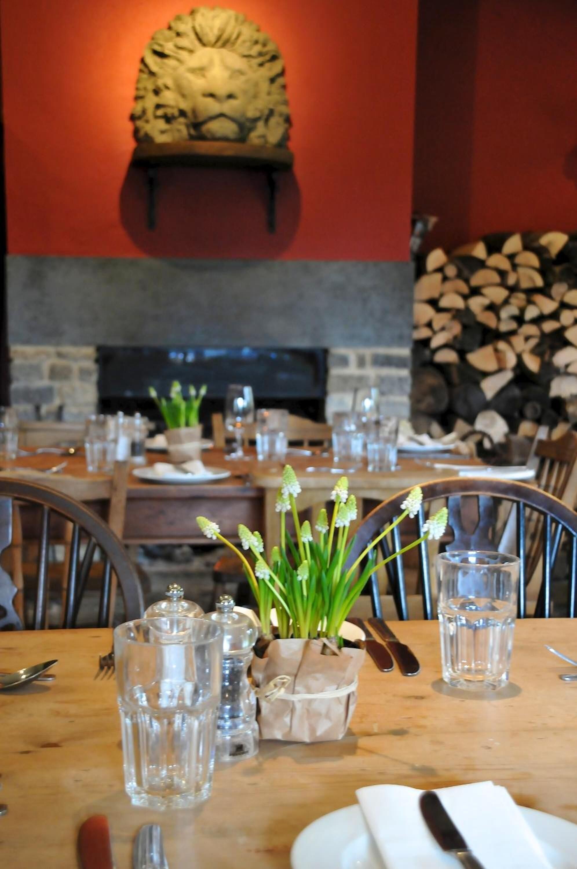 A restaurant interior with a fireplace, wooden tables, chairs, and a decorative lion head above the fireplace.