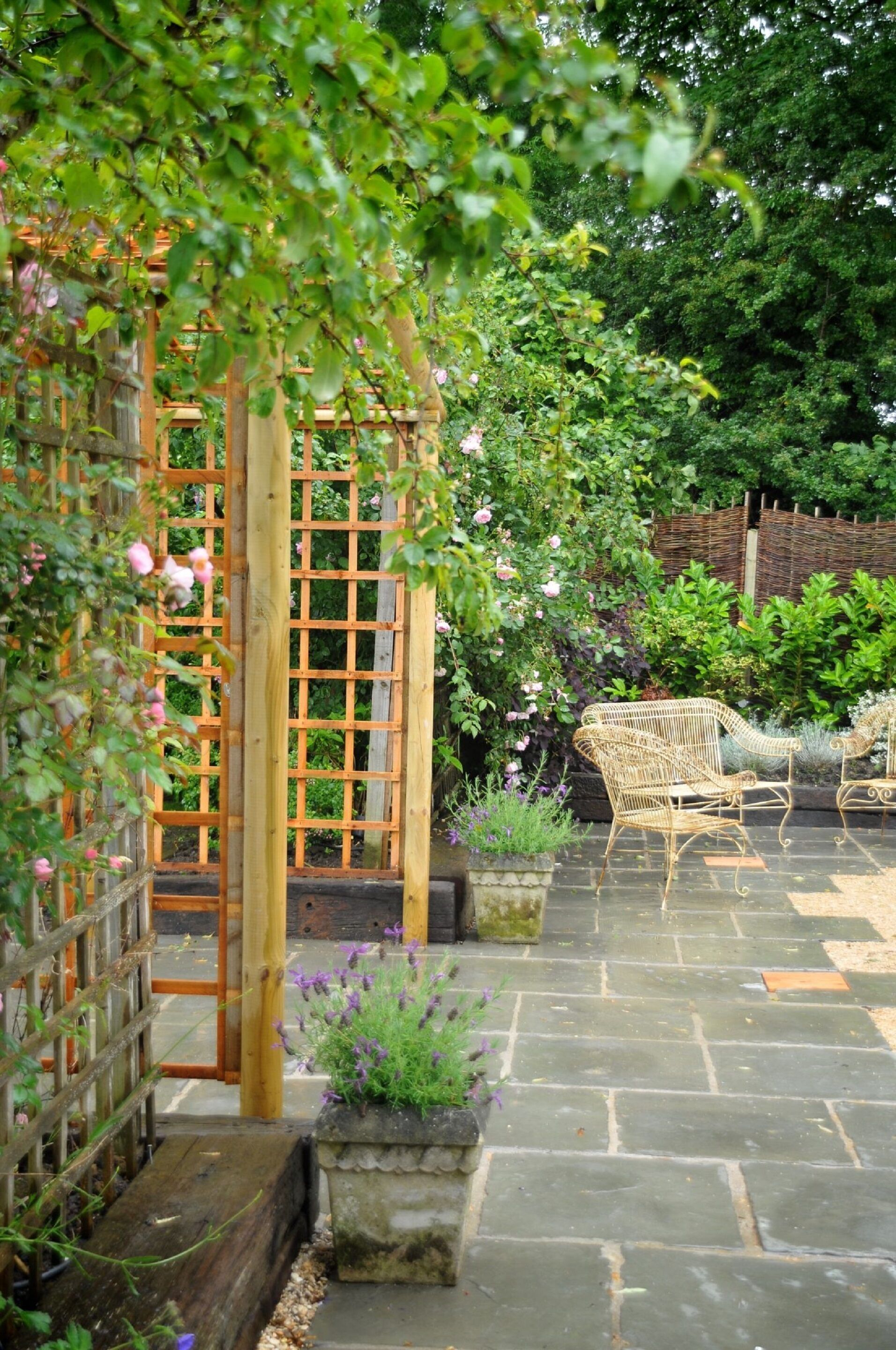 Une terrasse de jardin agrémentée d’une treille en bois, d’un sol en pierre et de meubles en osier.