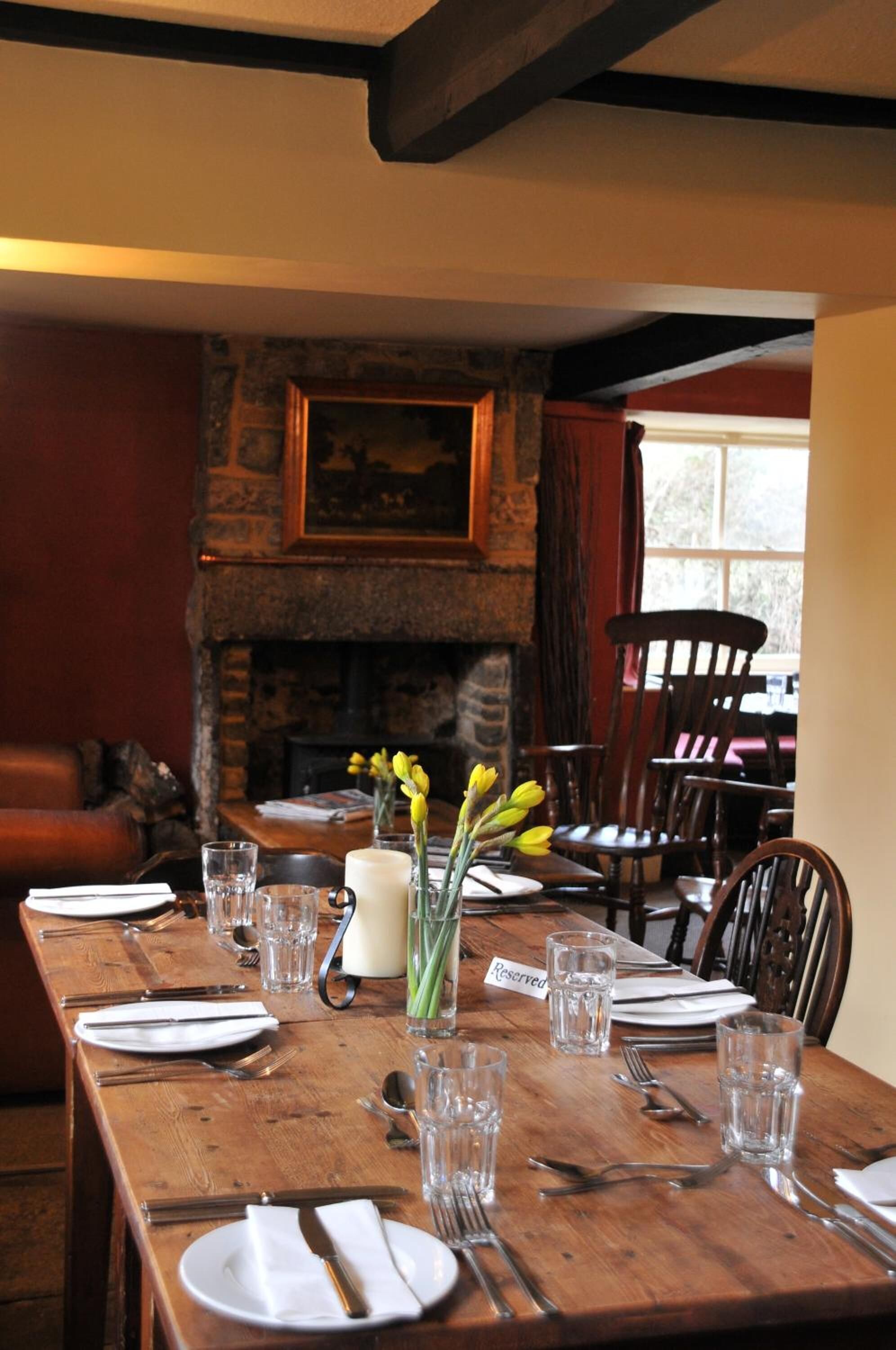 A wooden dining table set with plates, cutlery, and glasses, with a 'Reserved' sign, a vase with yellow flowers, and a stone fireplace in the background.