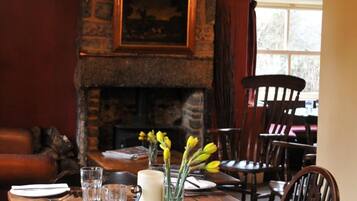 A wooden dining table set with plates, cutlery, and glasses, with a 'Reserved' sign, a vase with yellow flowers, and a stone fireplace in the background.
