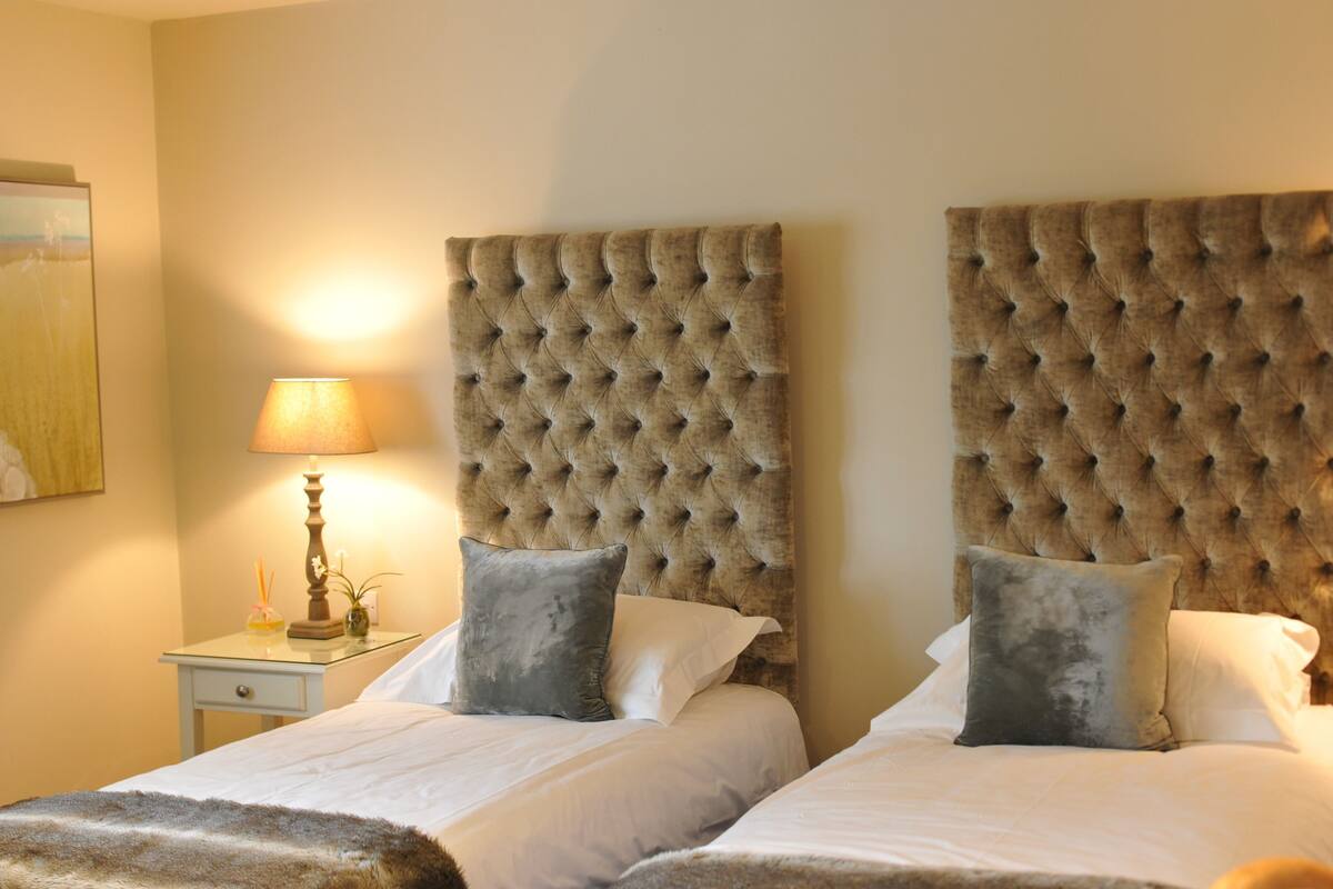 A hotel room with two beds, each with a tufted headboard, a bedside table with a lamp, and a small plant.