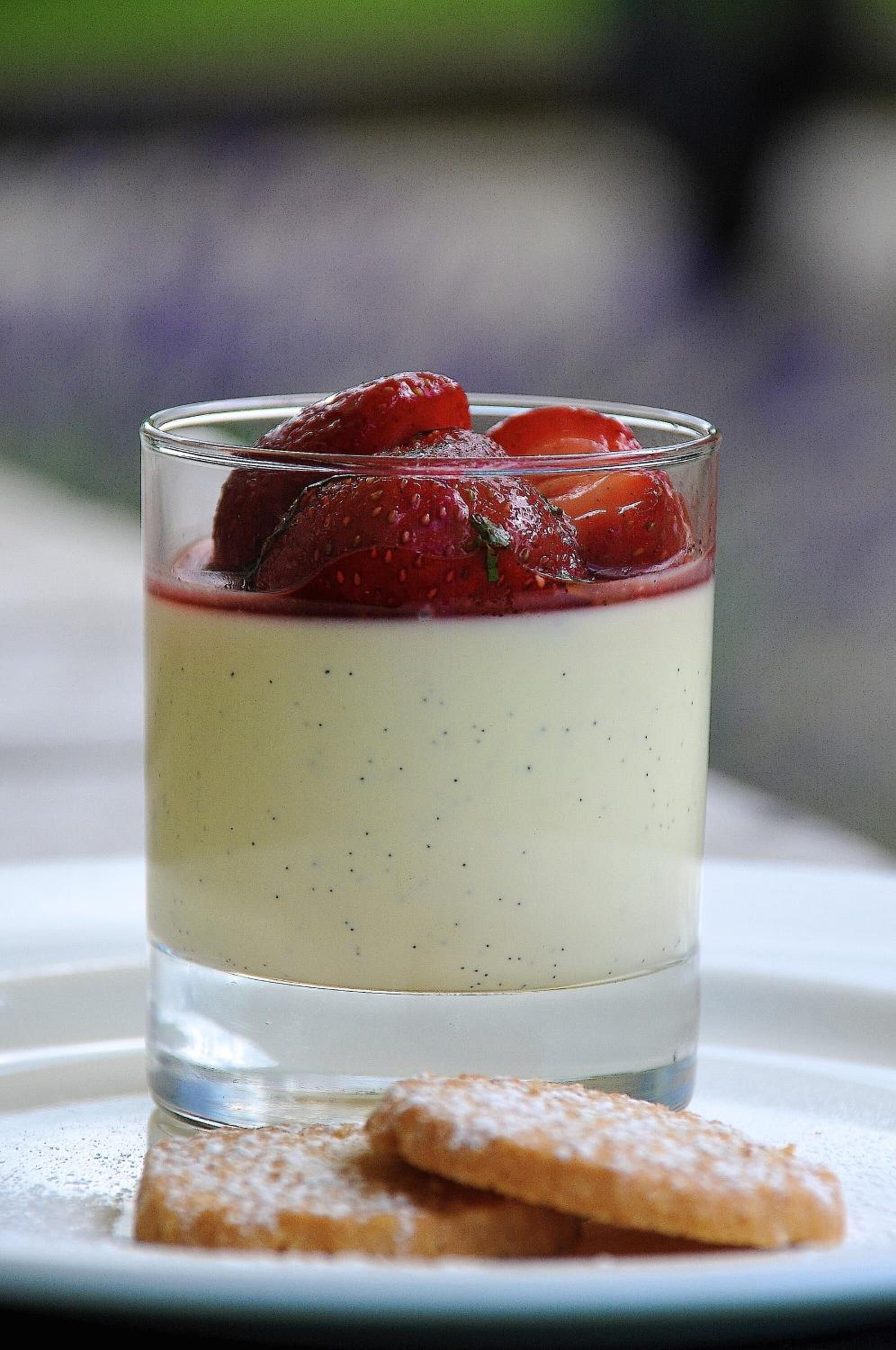 A dessert in a glass with strawberries on top, served on a plate with a biscuit.