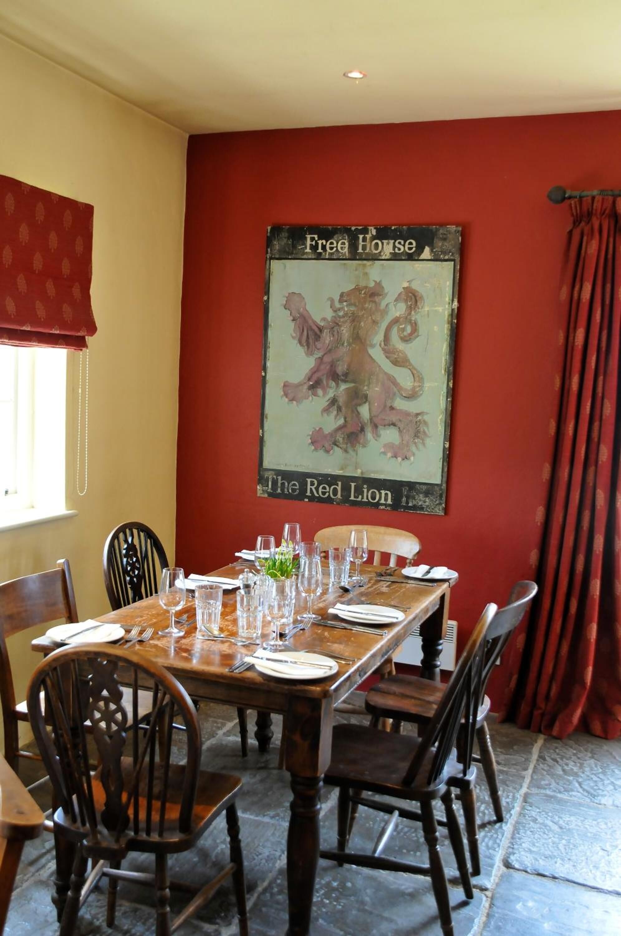 Ein Essbereich mit einem Holztisch, gedeckt für vier Personen, umgeben von Holzstühlen. Ein roter Vorhang und ein Fenster mit einem gemusterten Gardine sind sichtbar. Ein Schild an der Wand zeigt: "Free House The Red Lion".