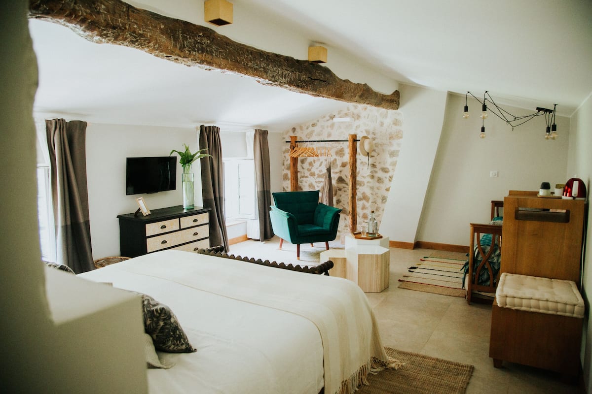 A bedroom with a bed, a green chair, a wooden dresser, a television, and a stone wall.