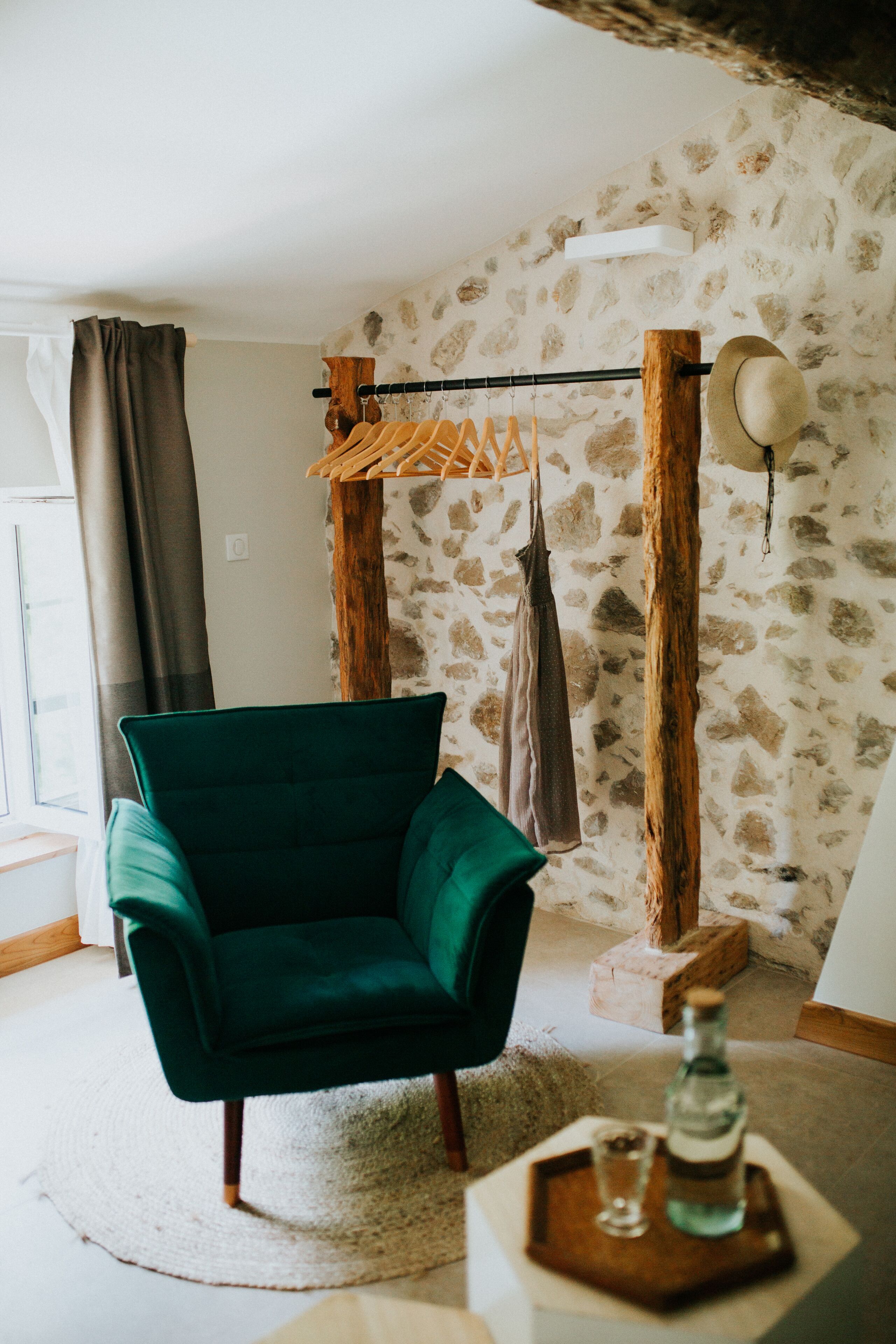 A room with a green armchair, a wooden coat rack, a stone wall, and a bottle on a tray.