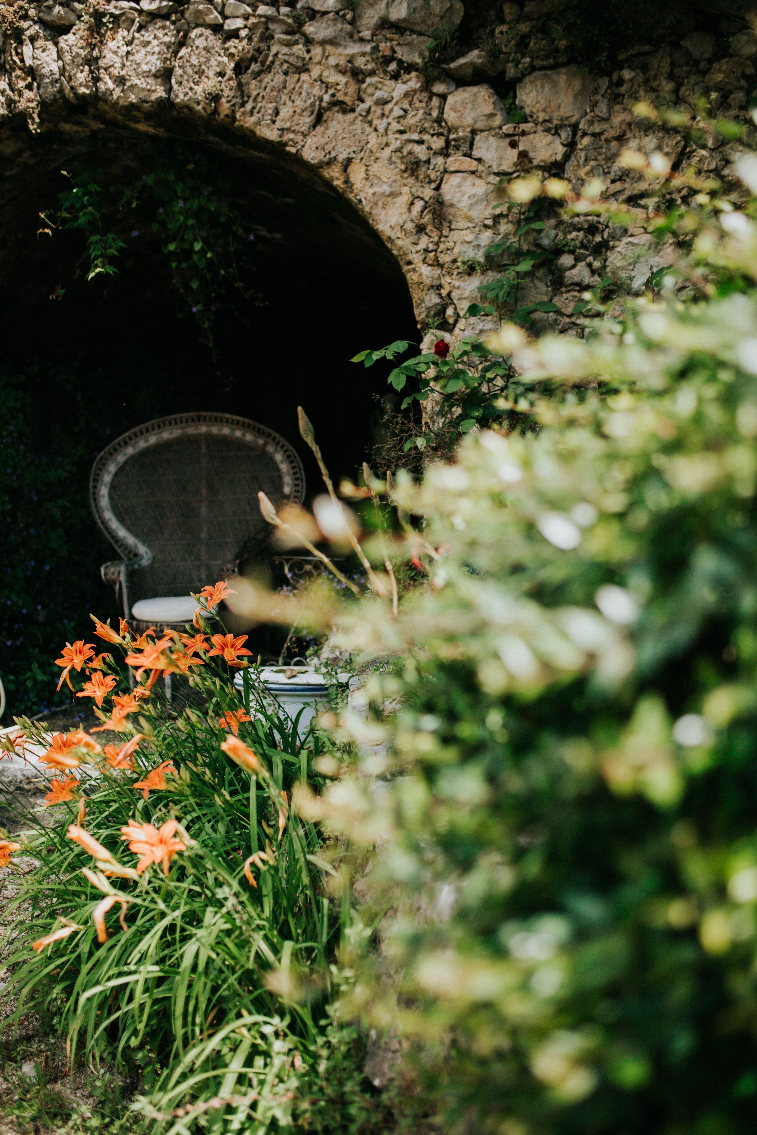 Un arco de piedra que da paso a un espacio con una silla y una mesa, rodeado de follaje y flores.
