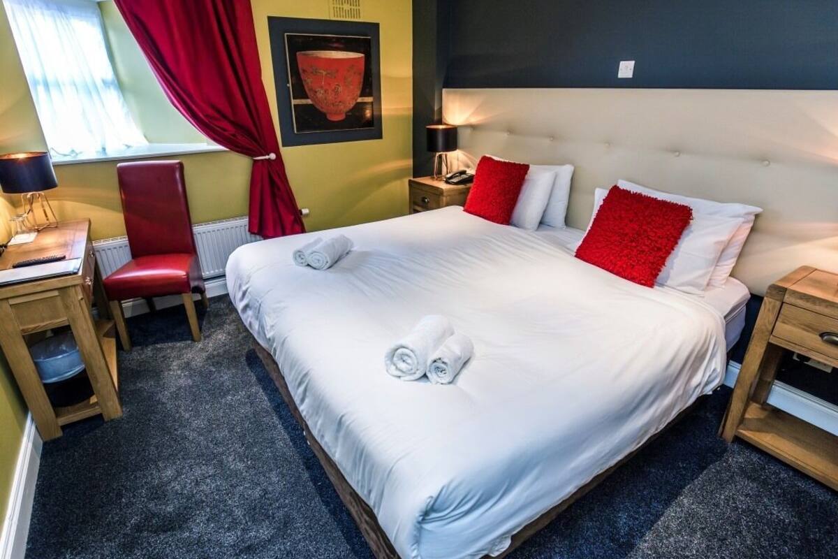 A neatly made bed with white linens and red pillows, a wooden nightstand with a lamp, a red chair, and a small table with a lamp.
