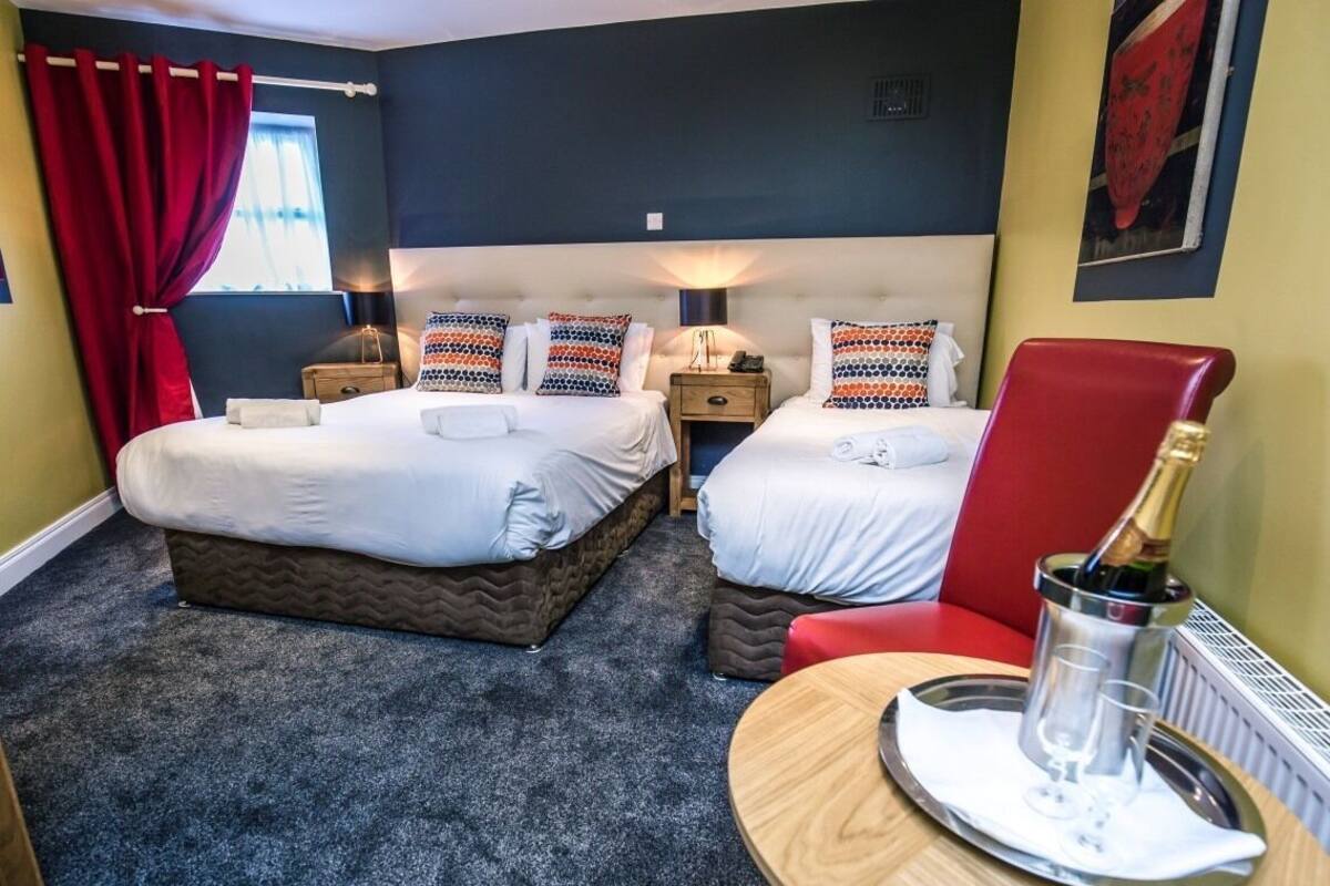 A hotel room with two beds, a red armchair, a bedside table with a lamp, and a tray with a bottle and glasses.