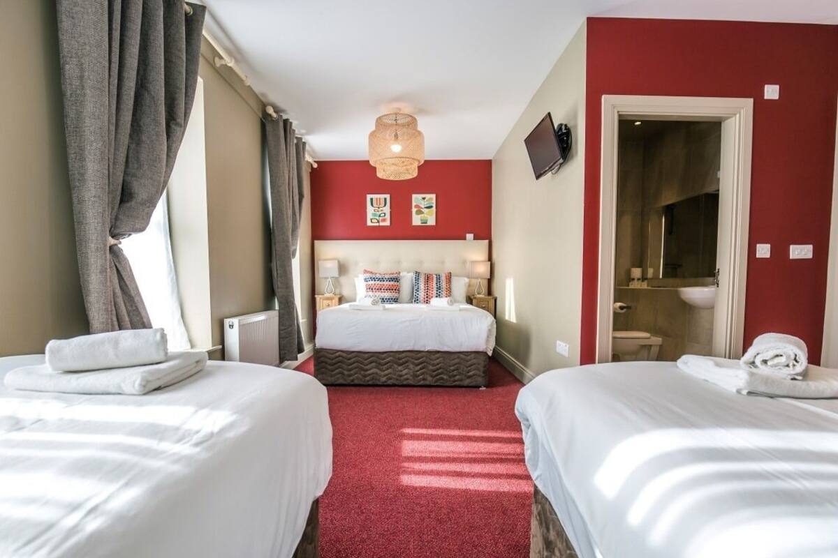 A hotel room with two beds, a red carpet, a large bed with multiple pillows, a television, and a bathroom visible through an open door.