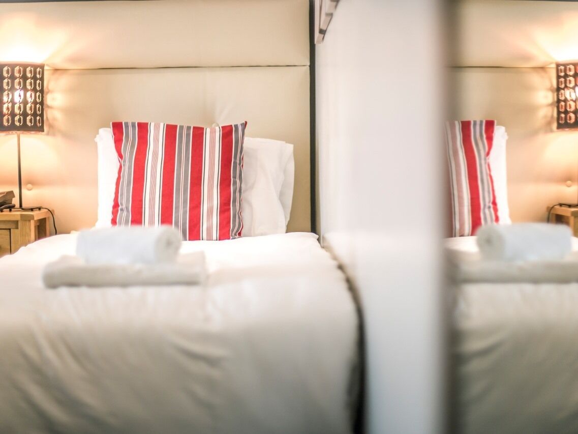 Habitación de hotel con dos camas, cada una con un cojín a rayas y una toalla enrollada. Hay lámparas de noche y una mesita de noche con una lámpara.