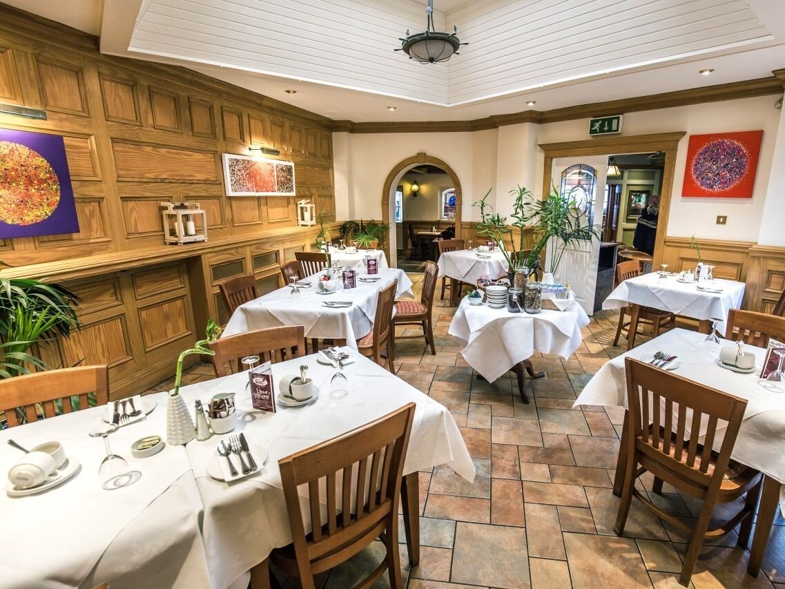 Ein Restaurant mit Holzwandverkleidung, weißen Tischdecken und Holzbänken. Die Tische sind mit Tellern, Besteck und Gläsern gedeckt.