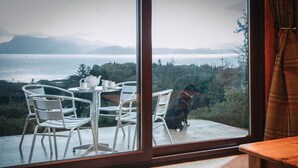 Een kamer met een groot raam dat uitzicht biedt op een prachtig uitzicht over een waterpartij en bergen. Binnenin staat een tafel met stoelen en zit er een hond naast het raam.