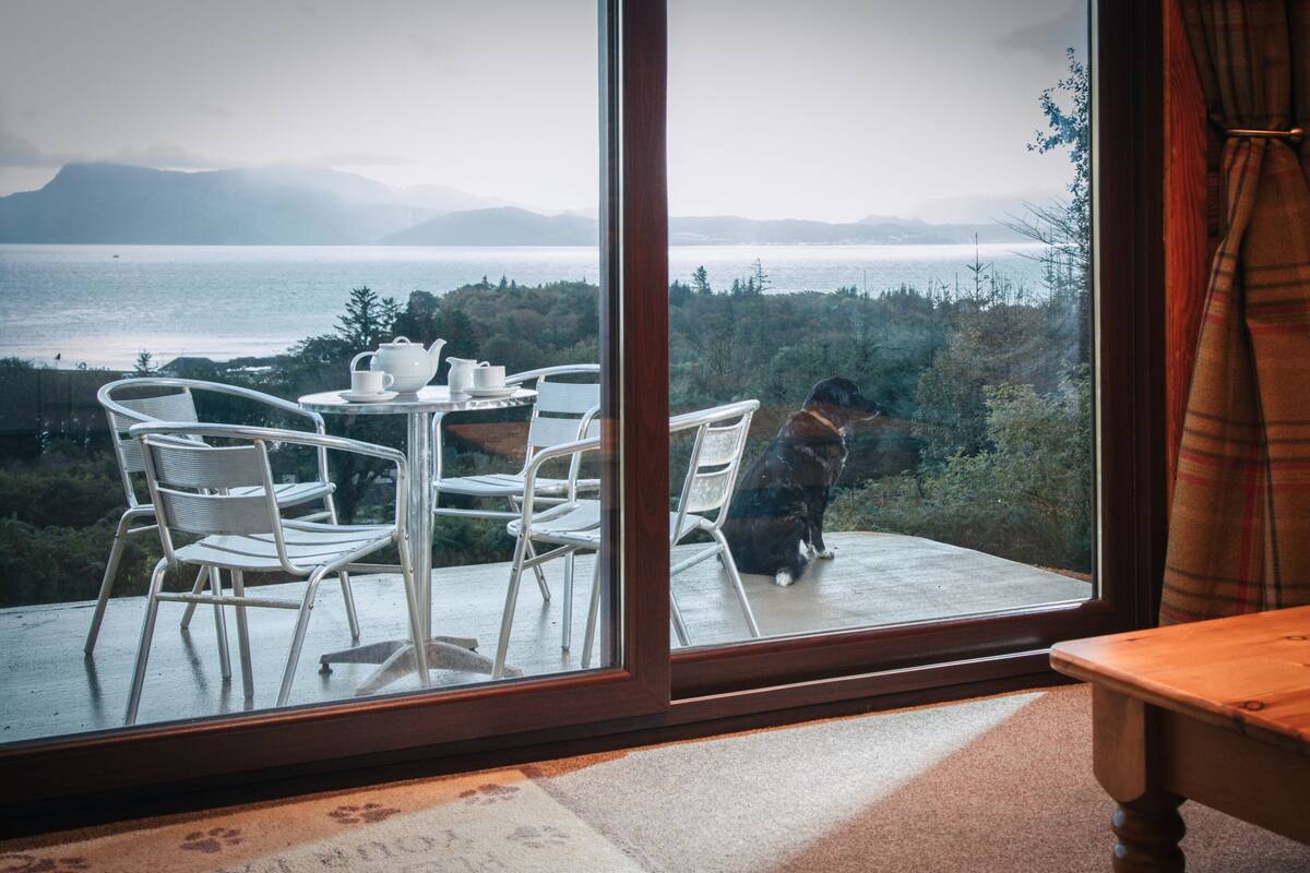 A room with a large window overlooking a scenic view of a body of water and mountains. Inside, there is a table with chairs and a dog sitting by the window.