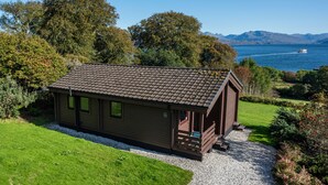 Een klein huisje met een schuin dak, omgeven door een verzorgde tuin en uitzicht op een meer met bergen op de achtergrond.