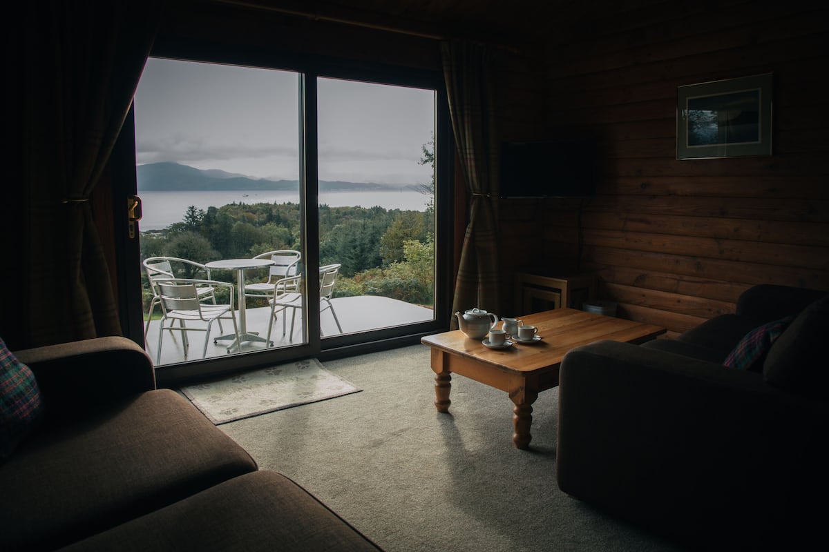 A cozy room with a large sliding glass door leading to a balcony. Inside, there is a wooden table with a teapot and cups, a sofa, and a chair.