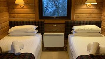 Ein Blockhaus-Zimmer mit zwei Einzelbetten, Karo-Bettwäsche, einem Fenster mit Blick auf Bäume und Wandlampen.